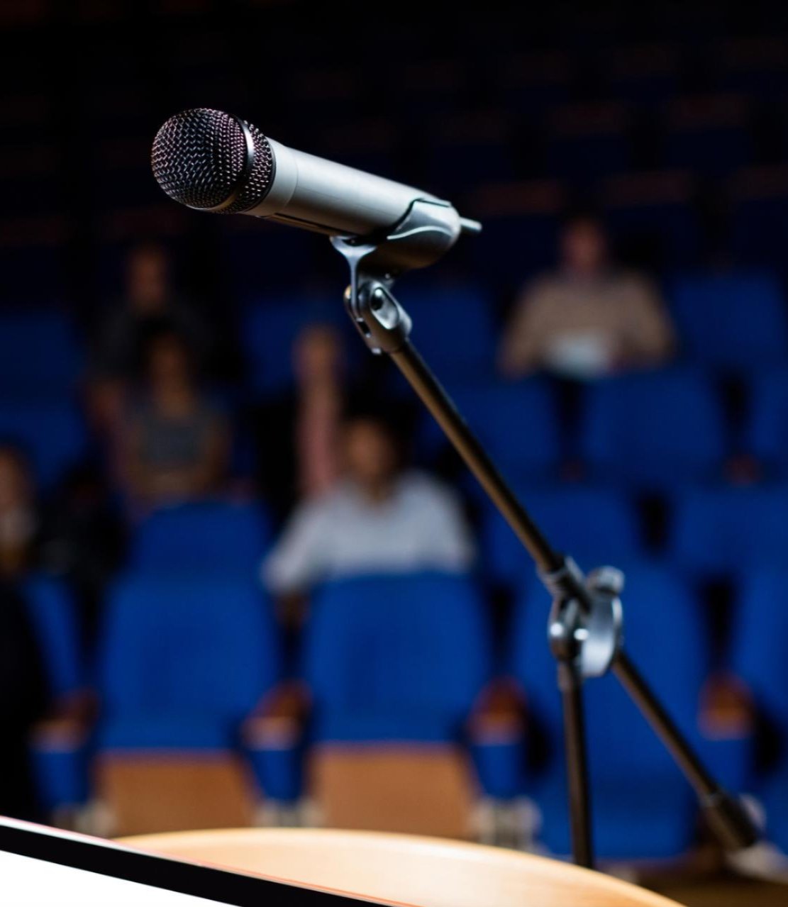 A close-up of a microphone mounted on a stand, positioned in front of an audience seated in blue chairs. The blurred background suggests a lecture hall or auditorium setting, emphasizing the microphone as the focal point.